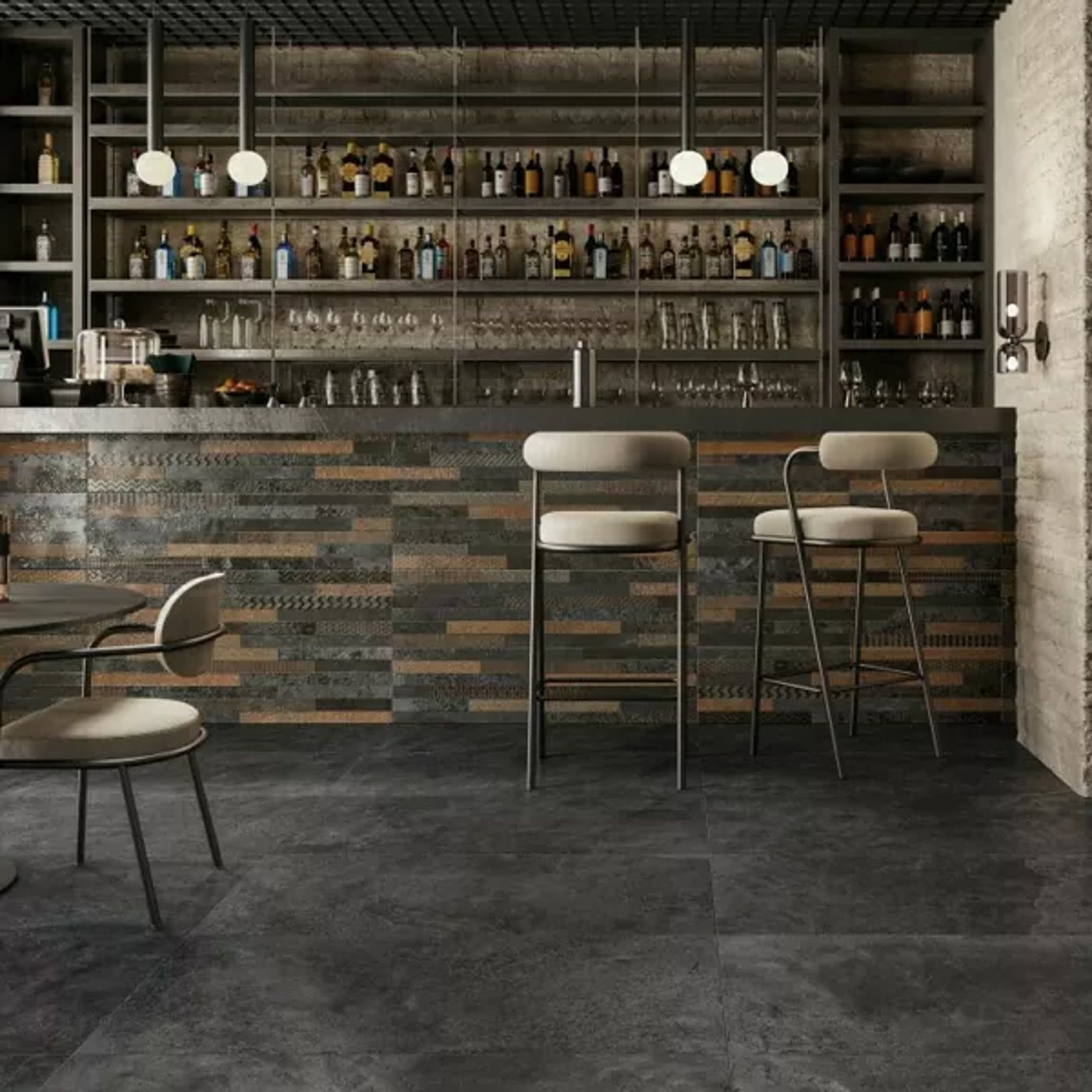 Intérieur de bar au comptoir habillé de carreaux effet pierre anthracite et cuivré, avec deux tabourets hauts crème, une étagère de bouteilles et un sol en carrelage gris foncé.