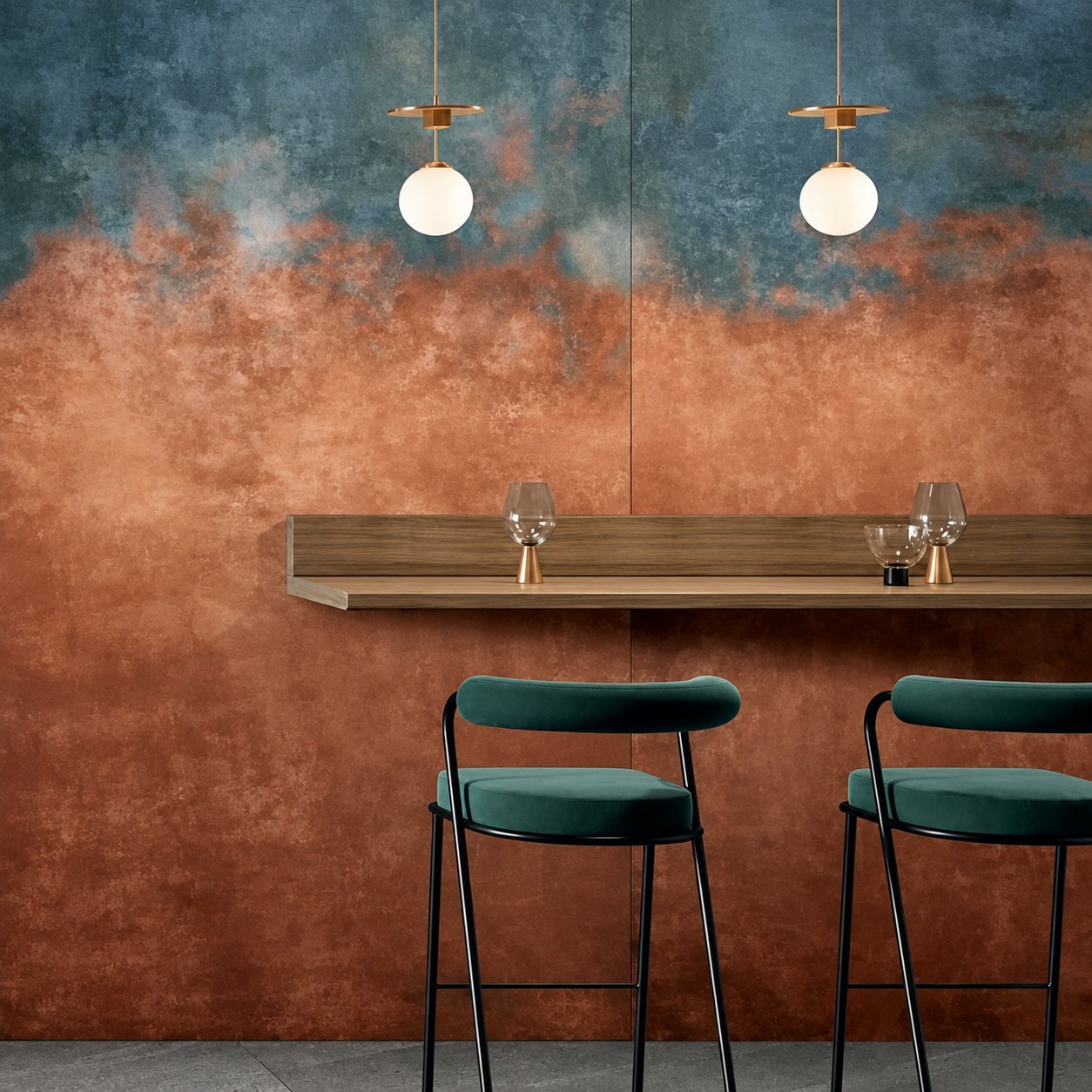 Deux tabourets de bar verts devant une tablette en bois fixée à un grand mur aux teintes cuivrées et bleutées, sous deux suspensions en forme de globe.