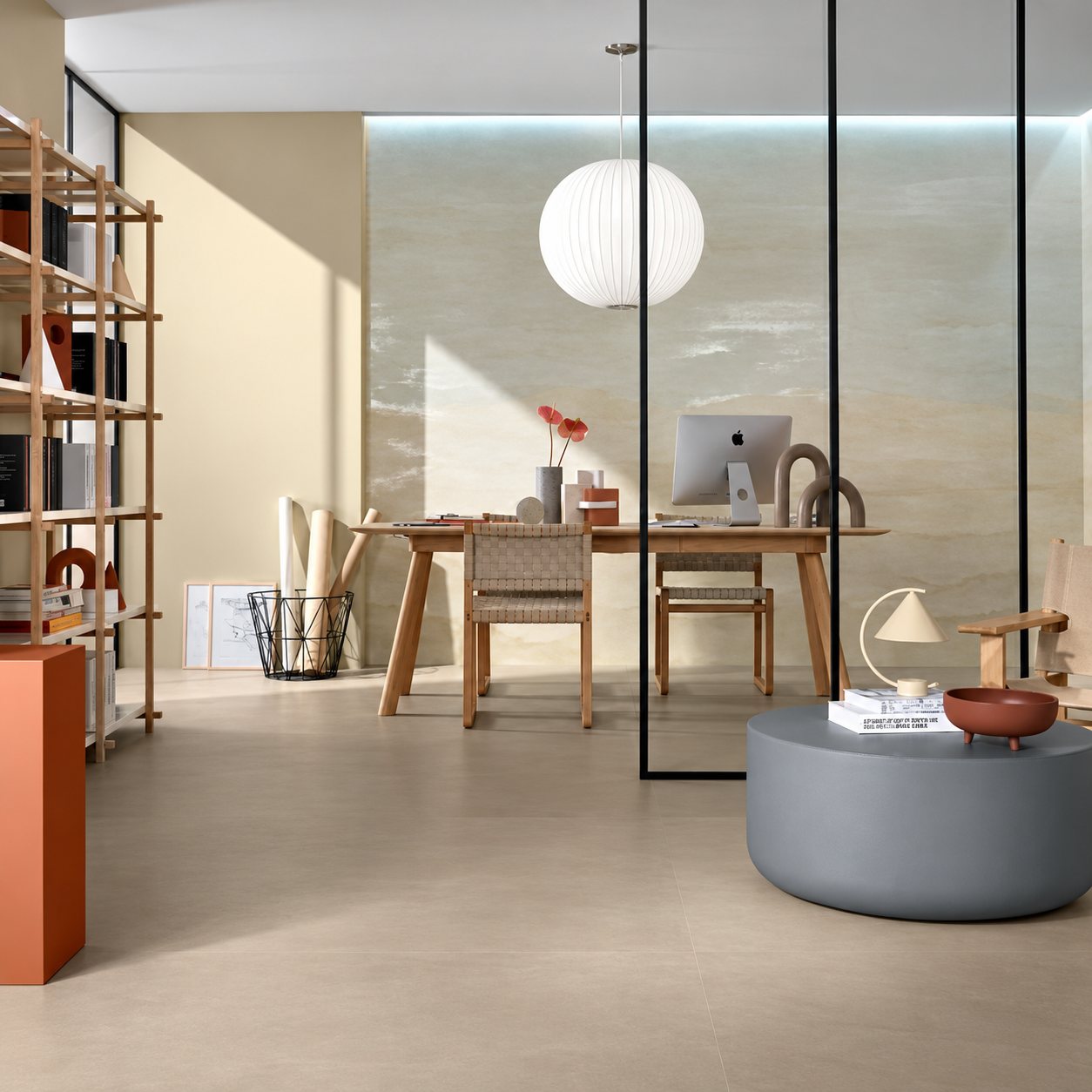 Un espace de bureau lumineux avec sol et mur en carrelage beige, table en bois, suspension blanche ronde, étagère et pouf gris séparés par une verrière noire.