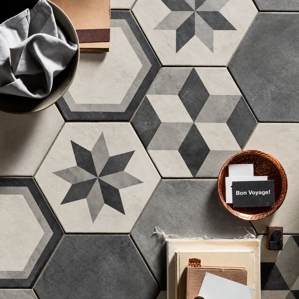 Vue de dessus de carreaux hexagonaux aux motifs d'étoiles et de cubes en gris et beige, avec un bol en cuivre, un tissu gris et des carnets posés dessus.