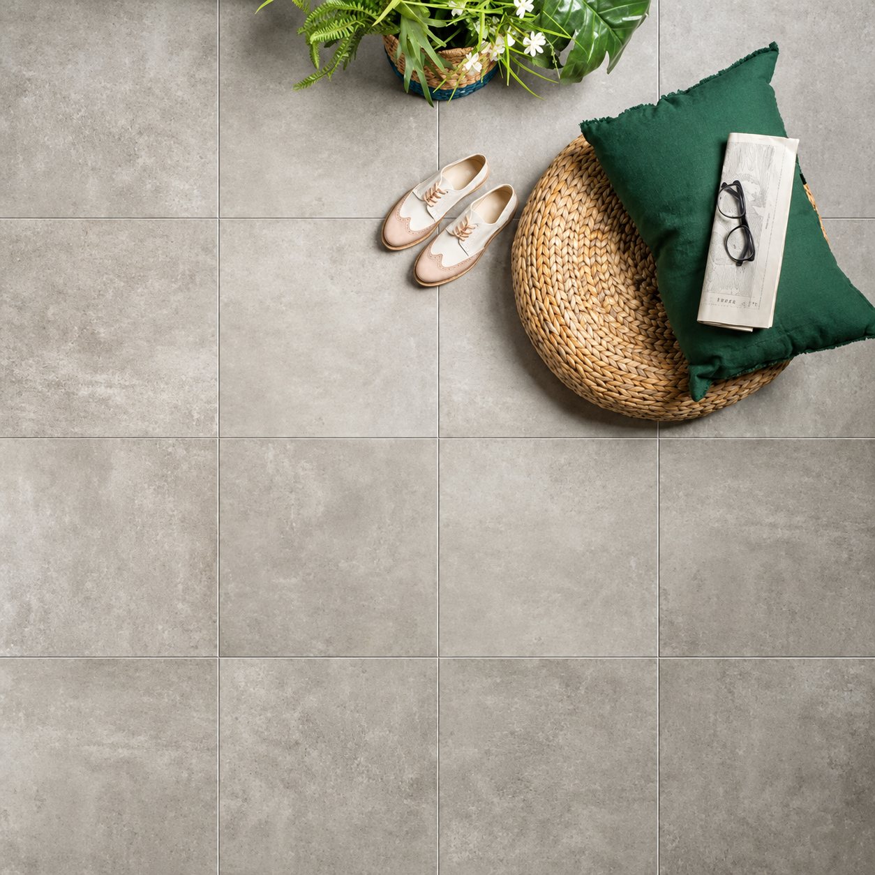 Vue de dessus d'un sol en grands carreaux gris effet béton décoré d'un pouf tressé, d'un coussin vert, d'un journal avec lunettes, de chaussures claires et d'une plante.