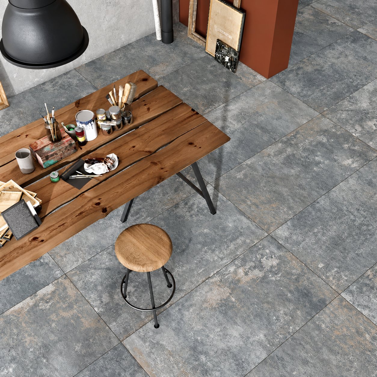 Vue de dessus d'un sol en grand carrelage gris effet pierre avec une table en bois brut couverte de pinceaux et de pots de peinture et un tabouret rond.