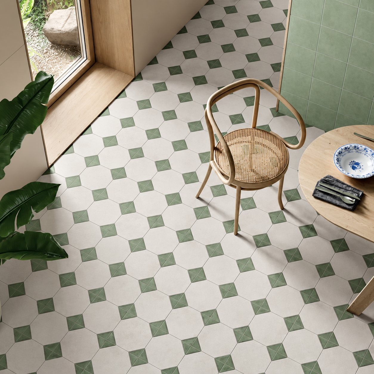 Vue en plongée d'un sol en carreaux octogonaux beiges à cabochons verts, avec une chaise en bois cannée, une table ronde et une plante.