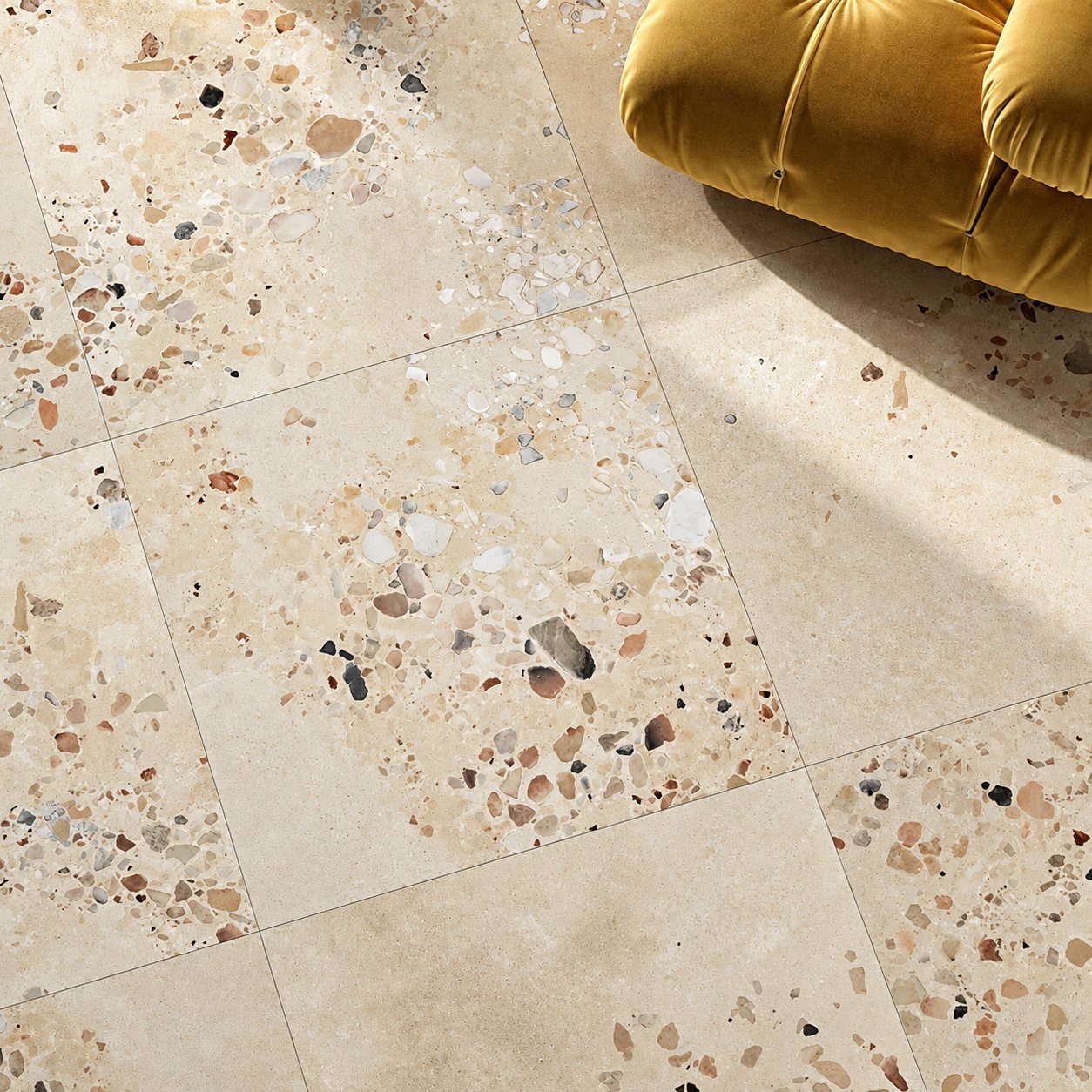 Sol en carrelage beige effet terrazzo à éclats colorés, avec l'angle d'un canapé en velours jaune moutarde et des ombres portées.