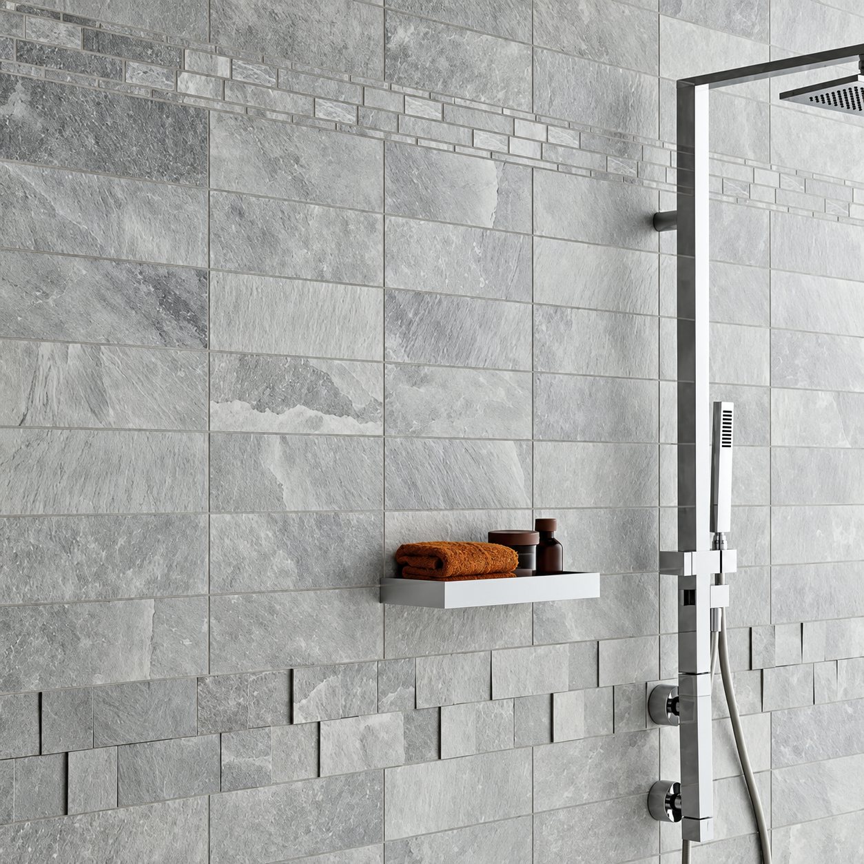 Mur de douche habillé de carrelage gris effet pierre avec une colonne de douche chromée et une petite étagère portant une serviette orange.