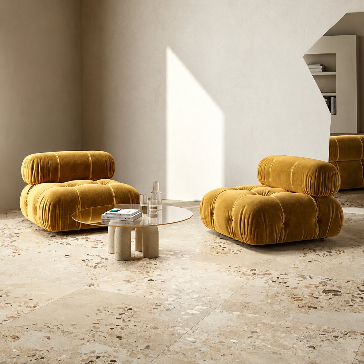 Deux fauteuils modulaires capitonnés en velours jaune moutarde entourent une table basse ronde en verre posée sur un sol en terrazzo beige, dans un salon lumineux.