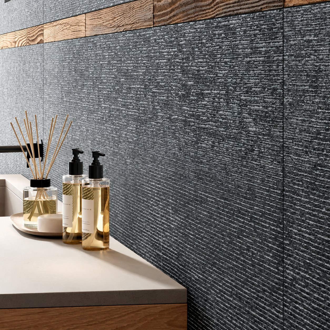 Mur de salle de bain habillé de carreaux gris anthracite texturés et de bandes de bois, avec un diffuseur de parfum à bâtonnets et deux flacons de savon posés sur un plan beige.
