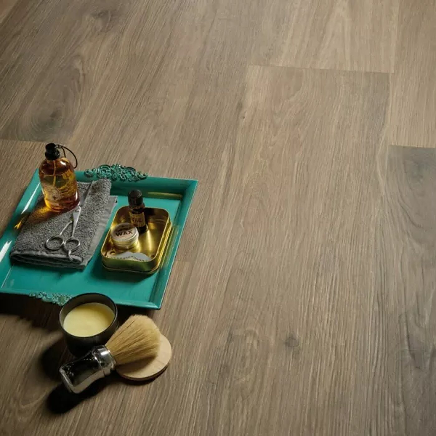Un plateau turquoise orné avec ciseaux, serviette grise, flacon d'huile, boîte de cire et blaireau de rasage est posé sur un sol imitation parquet brun clair.