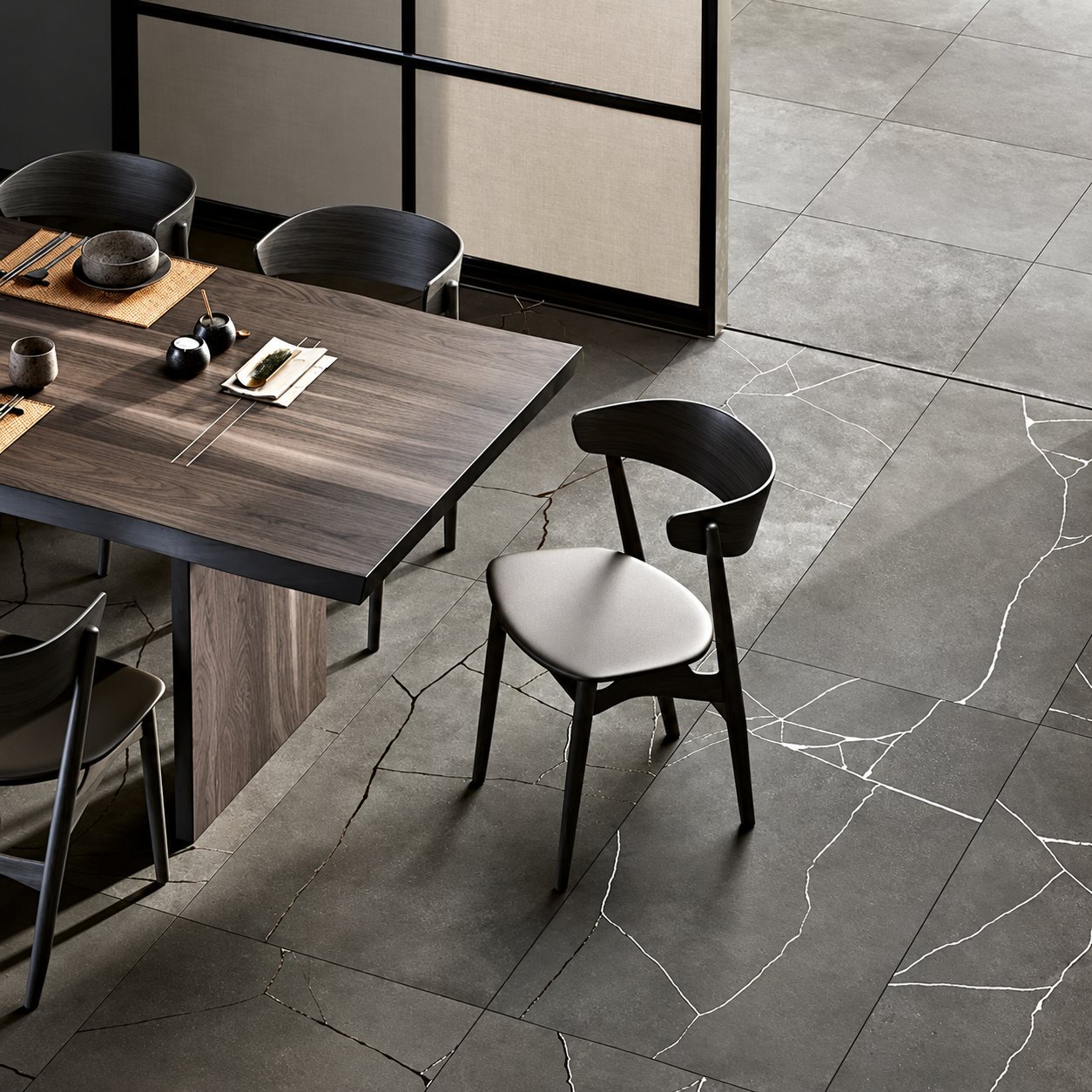 Vue en plongée d'une table en bois sombre et de chaises noires sur un grand carrelage gris foncé aux veines blanches fines.