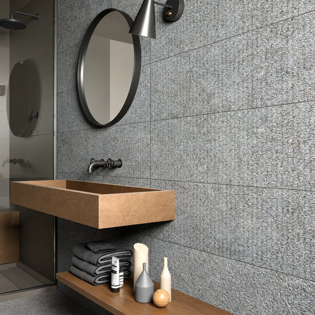 Salle de bain contemporaine aux murs en carrelage gris texturé, avec un miroir rond, une vasque rectangulaire en bois et des serviettes grises pliées sur une étagère.