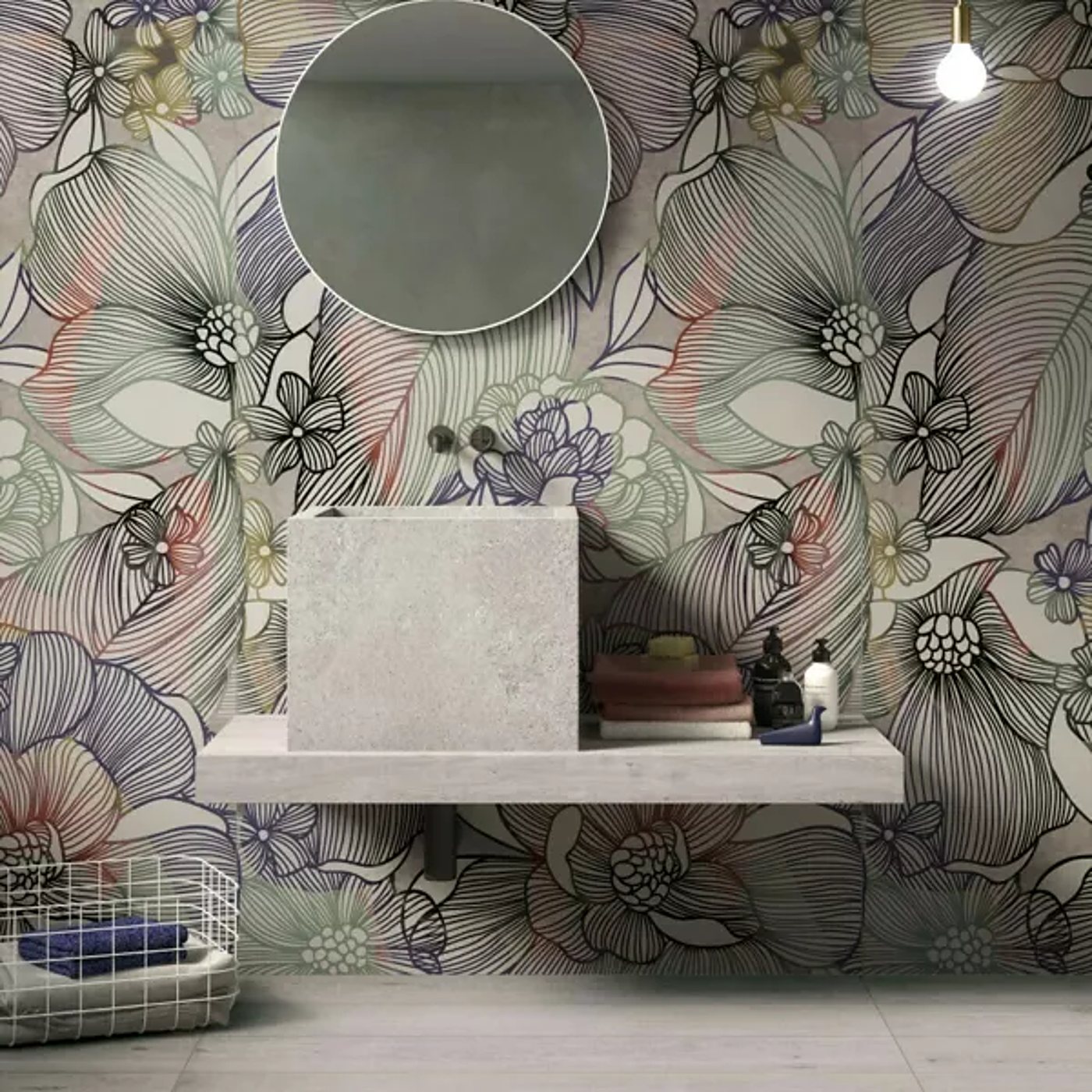 Salle de bains au papier peint à grandes fleurs colorées, avec un miroir rond, une vasque carrée en pierre grise posée sur une tablette et un panier en fil blanc.