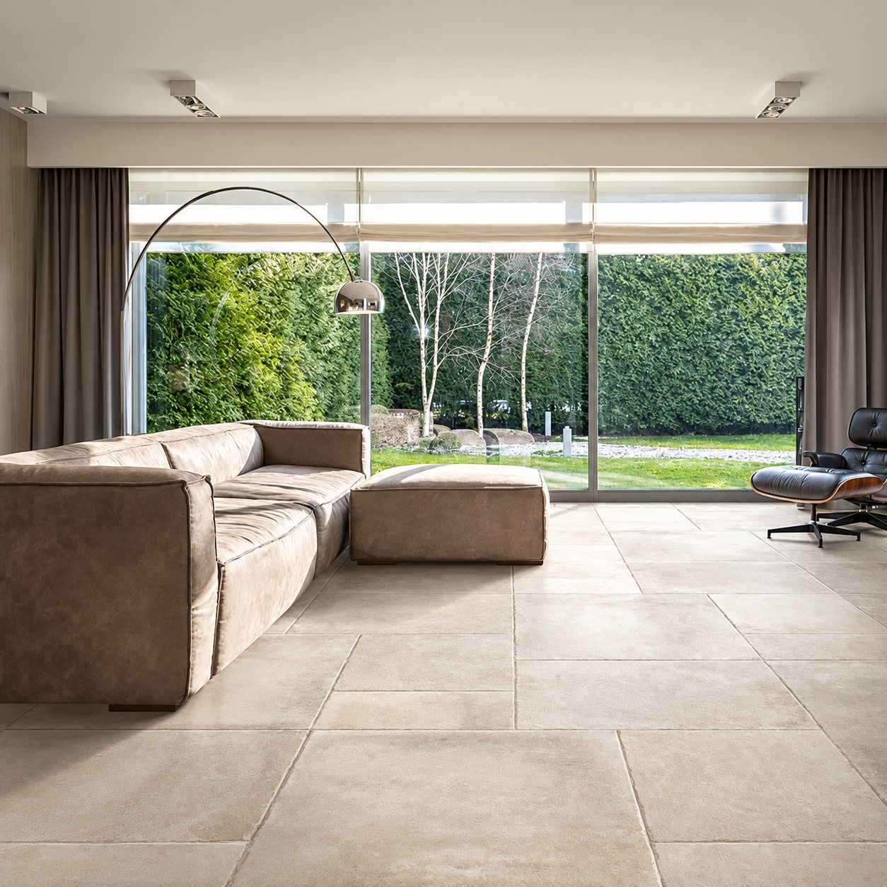 Salon lumineux au sol en grands carreaux beige clair, avec canapé en cuir, lampadaire arqué et grande baie vitrée donnant sur un jardin.