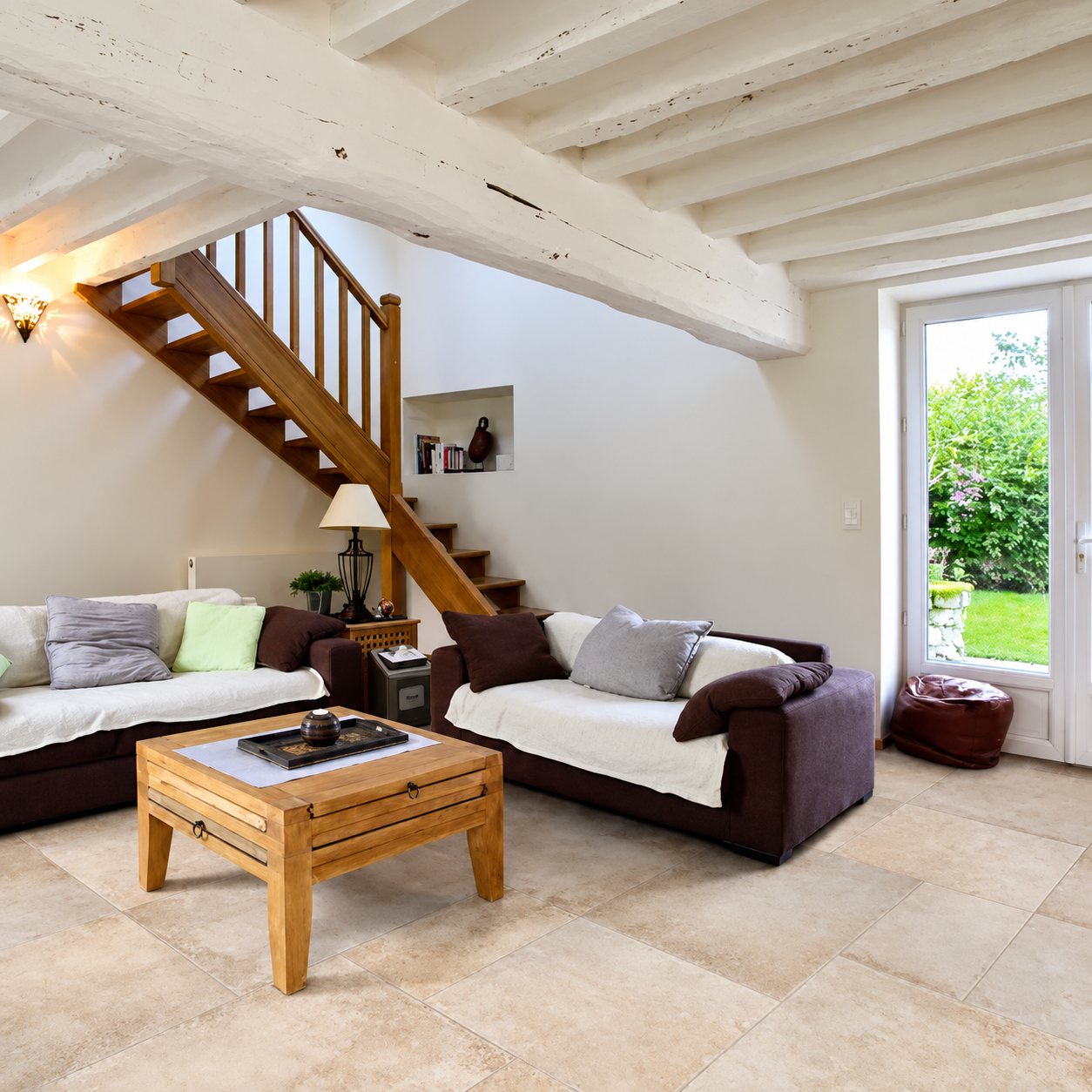 Salon clair aux poutres apparentes blanchies, avec deux canapés bruns, une table basse en bois, un escalier et un sol en grand carrelage effet pierre.
