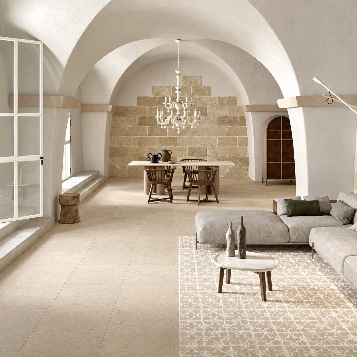 Un vaste salon aux voûtes blanches, avec un sol en carrelage beige, un canapé gris, une table à manger et un lustre blanc.