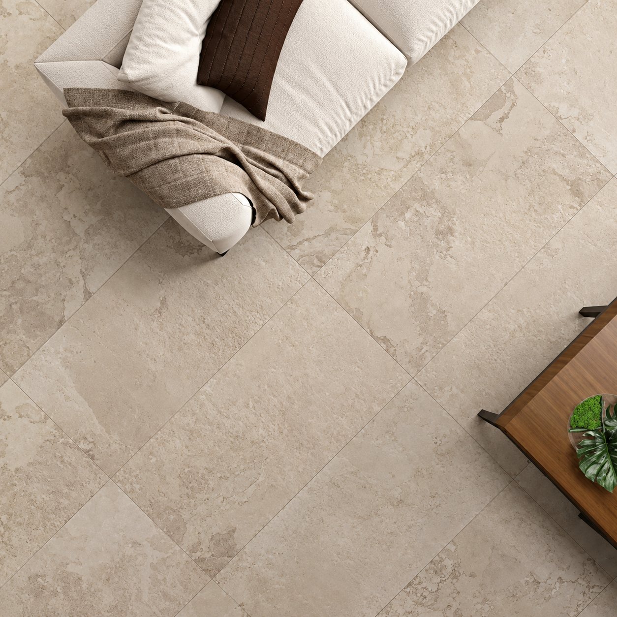 Vue de dessus d'un grand carrelage beige effet pierre posé en diagonale, avec un canapé crème, un plaid marron et une table basse en bois.